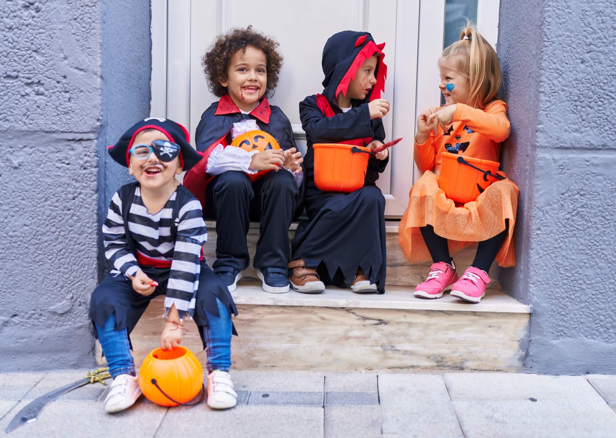 Four children in Halloween costumes, including a pirate, a vampire, and a pumpkin, sit on steps holding candy buckets.