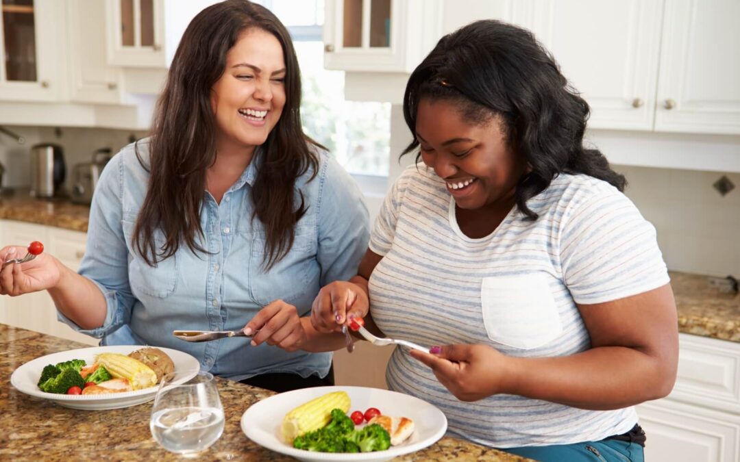 Two smiling women in a bright kitchen share a healthy meal of chicken, corn, and broccoli on plates set on a granite countertop.